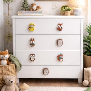 May include: A white dresser with six animal-themed drawer pulls. The pulls feature a bear in a yellow raincoat, a fox, a deer, a hedgehog, an owl, and a squirrel. The dresser is in a child's room with a forest-themed painting and stuffed animals.