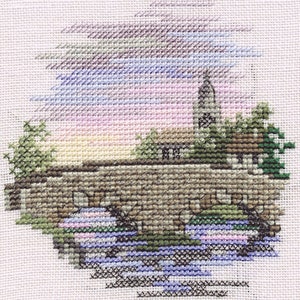 Peut inclure: Broderie au point de croix représentant une scène de village avec un clocher d'église, des maisons, un pont et une rivière. Le ciel est un dégradé de rose, violet et bleu.