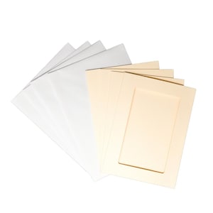 Puede incluir: Un conjunto de cartones de passe-partout y cartones de fondo blancos y crema, abanicados sobre un fondo blanco. Los passe-partouts tienen una abertura rectangular y los cartones de fondo son macizos.