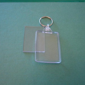 Puede incluir: Un llavero rectangular transparente con un anillo dorado. El llavero tiene un inserto de plástico transparente y una pieza de plástico transparente separada. El fondo es verde liso y apagado.