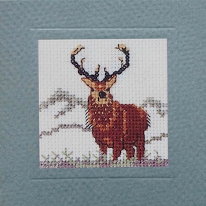 May include: A cross-stitch design of a brown deer with large antlers standing in a snowy field. The deer is facing forward with its head slightly tilted. The background is a white snowy field with some green bushes.