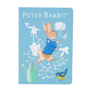 May include: A light blue Peter Rabbit journal with a diamond painting design. The cover features Peter Rabbit in a blue jacket, a blue bird, a watering can, and the text "Peter Rabbit".