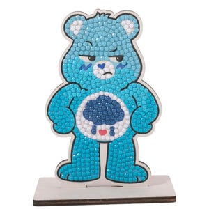 May include: A blue Care Bear ornament with a grumpy expression, decorated with sparkling blue gems. The bear has a cloud and rain design on its belly, with a red heart. It stands on a small wooden base, ideal for display.