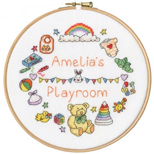 Op de afbeelding: Een witte borduurring met een kleurrijk kruissteekontwerp met een regenboog, wolken, sterren en verschillende speelgoedjes. De tekst "Amelia's Playroom" is in oranje geborduurd.