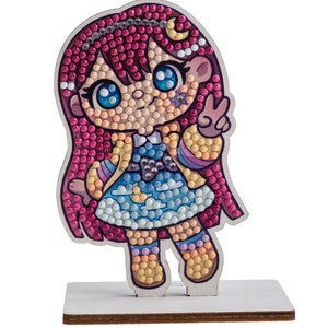 Peut inclure: Un kit d'artisanat en bois représentant une fille de dessin animé avec des cheveux roses, une veste bleue et jaune et une robe bleue avec des nuages et des étoiles. La fille lève deux doigts en signe de paix.