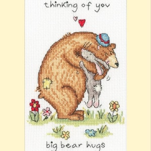 May include: A cross-stitch pattern featuring a brown bear wearing a blue hat hugging a grey rabbit. The text "thinking of you" is above the bear and rabbit, and the text "big bear hugs" is below them.