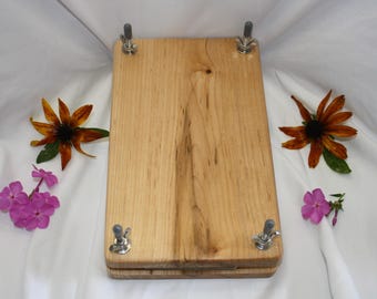 May include: A rectangular wooden flower press featuring a light brown wood grain finish. The device consists of two stacked wooden boards secured at each corner by metal bolts with silver wing nuts and grey plastic caps on the threaded ends.
