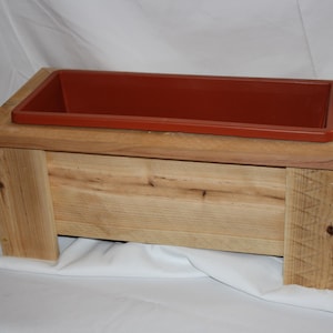 May include: A rectangular wooden planter box with a reddish-brown plastic insert. The planter is made of light-colored wood and has a simple, rustic design. The box is elevated on four sturdy legs.