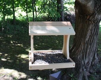 Può includere: Una mangiatoia per uccelli in legno chiaro con tetto spiovente e vassoio quadrato riempito di semi di girasole neri. La mangiatoia è sospesa tramite un sottile filo nero e presenta due supporti laterali curvi che collegano il tetto alla base.