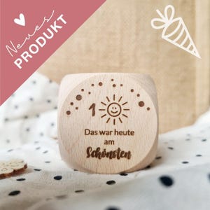 Könnte beinhalten: Ein Holzblock mit graviertem Text und einem Sonnendesign. Der Block hat die Zahl 1 mit einem lächelnden Sonnengesicht, und die Worte "Das war heute am Schönsten" stehen darunter. Der Hintergrund umfasst ein Tupfenmuster und ein rosa Banner mit den Worten "Neues Produkt".