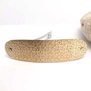 May include: A gold-colored hair barrette with an intricate geometric pattern. The barrette has a rectangular shape with rounded ends and a silver-colored clasp. The surface is adorned with a repeating design of squares and floral shapes.