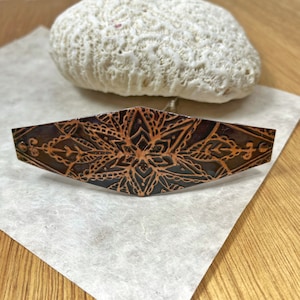 May include: A copper-colored hair barrette with an intricate floral and geometric design. The barrette is approximately 4 inches long and features a detailed, embossed pattern. The background includes a white, textured stone and a piece of paper.