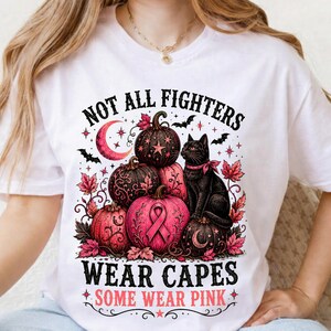 May include: A white t-shirt featuring a graphic print of pumpkins, a black cat with a pink ribbon, and a pink crescent moon. The text reads: NOT ALL FIGHTERS WEAR CAPES SOME WEAR PINK. The design includes pink autumn leaves, stars, and bats.