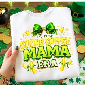 May include: White t-shirt with a St. Patrick's Day theme. The shirt features the text "in my STRING CHEESE MAMA ERA" in green and yellow, with a green bow and gold accents. The shirt is held by a person with green nail polish.