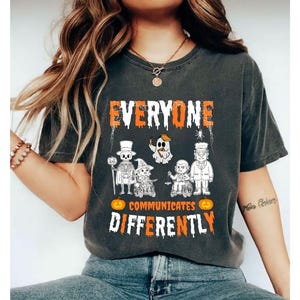 May include: Dark gray t-shirt with the text "EVERYONE COMMUNICATES DIFFERENTLY" in orange and white. The shirt features Halloween-themed characters, including a skeleton, ghost, and witch, with pumpkins and spiderweb details.