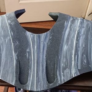 May include: A grey and white foam costume piece with a textured surface. The piece is shaped like a chest plate and has a curved design.