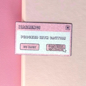 May include: A pink sticker resembling a computer pop-up window with the text "WARNING! PROCEED WITH CAUTION." It features "GO BACK" and "CONTINUE ANYWAYS" buttons, and a cursor. The sticker is set against a pink background.