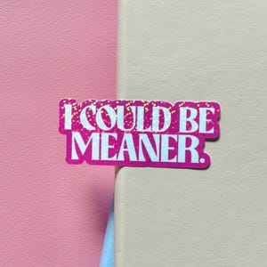 May include: A die-cut sticker featuring the text I COULD BE MEANER in bold, light blue, sans-serif capital letters. The background of the text is a vibrant magenta color with a subtle, scattered gold glitter or confetti-like texture throughout.