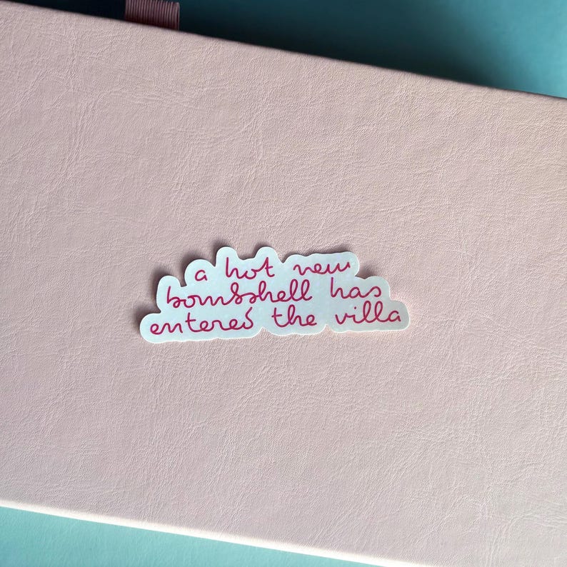 May include: A sticker with the phrase "a hot new bombshell has entered the villa" in pink script. The sticker is white with a cloud-like shape and is placed on a pink surface.