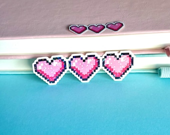 May include: Three pixelated pink heart stickers with white borders are arranged on a light pink notebook. The hearts have a glittery effect. Above them, three smaller heart stickers are also visible. The background is a light blue surface.