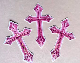 May include: Three pink cross stickers with white outlines and glitter accents. The crosses have a decorative, ornate design. The background is a solid, neutral color.