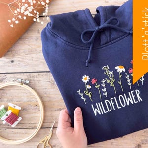 Puede incluir: Una sudadera con capucha azul marino con un diseño bordado en blanco de flores silvestres y la palabra "WILD FLOWER". La sudadera tiene una capucha con cordón y un bolsillo canguro.