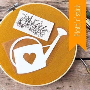 Op de afbeelding: Een witte gieterstencil met een hartvormige uitsparing op een gele stoffen achtergrond. De stencil ligt op een stuk borduurring. Een klein stukje witte stof met een zwart bloemenborduurpatroon is ook zichtbaar.