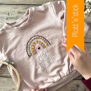 Può includere: Una felpa rosa con un arcobaleno ricamato bianco e il testo "little sunshine".