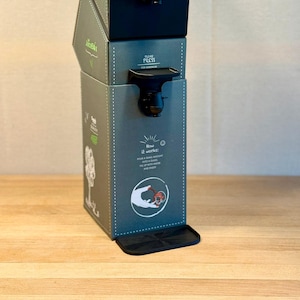 Peut inclure: Trois distributeurs JuiceBox empilés, verts et noirs. Chaque boîte a un robinet noir et les mots "PLEASE PRESS" et "JuiceBox". La boîte du bas contient des instructions et un graphique expliquant comment utiliser le distributeur.