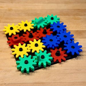 Può includere: Un puzzle colorato con ingranaggi in plastica ad incastro gialli, verdi, rossi e blu. Gli ingranaggi sono disposti su una base nera, creando un giocattolo visivamente accattivante ed educativo. Lo sfondo è una superficie in legno.