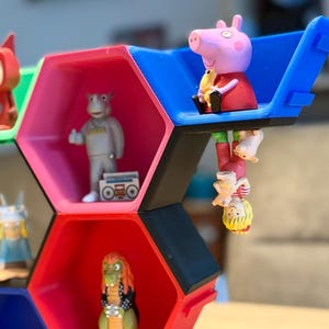 Può includere: Una colorata mensola per giocattoli geometrica con scomparti esagonali, ognuno dei quali ospita una diversa statuina. La mensola è principalmente rossa, blu e nera, con altri colori. Le statuine includono un maiale dei cartoni animati, un ippopotamo con un boombox e altri personaggi.