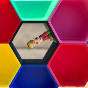 Könnte beinhalten: Buntes, geometrisches Regal mit sechseckigen Fächern in Rot, Grün, Gelb, Blau und Pink. Eine kleine Figur einer Person in roter und grüner Kleidung befindet sich in einem der Fächer.