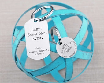 May include: A silver keychain with two tags. The larger tag reads "BEST. Bonus DAD. EVER." and the smaller tag reads "BEST. Bonus DAD. EVER." with the names "emma audrey willia".
