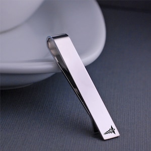 May include: A silver-colored tie clip with a curved top and a rectangular body. The clip has a small, black emblem near the bottom. The tie clip is angled, resting on a white surface and a gray background.