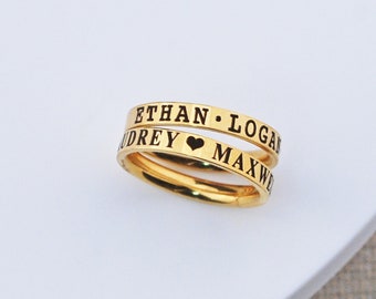 Puede incluir: Dos anillos de oro con nombres grabados en negro y un símbolo de corazón. Los anillos están apilados uno encima del otro. Los nombres en los anillos son "Ethan. Logan" y "Audrey. Maxwell".