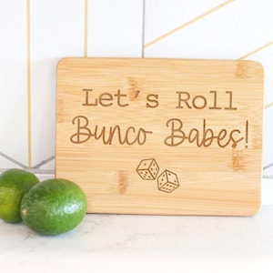 Peut inclure: Une planche à découper en bois avec le texte "Let's Roll Bunco Babes!" et une paire de dés gravés dessus.