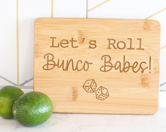 May include: A wooden chopping board with the text "Let's Roll Bunco Babes!" and a pair of dice engraved on it.