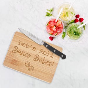 May include: A small wooden cutting board with the text "Let's Roll Bunco Babes!" engraved on it. A small paring knife is on the board. Two dice are also engraved on the board.