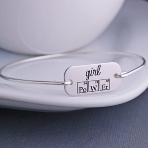 May include: Silver bangle bracelet with a rectangular tag that reads "girl" and features the chemical symbols for Polonium, Tungsten, and Erbium, with their atomic numbers.