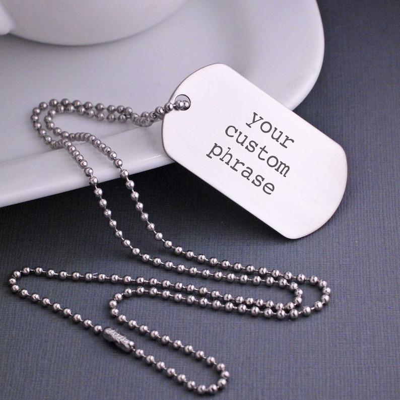 May include: A silver dog tag necklace with a ball chain. The dog tag has the text "your custom phrase" engraved on it.