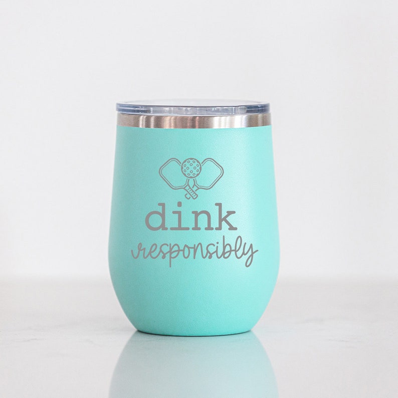 May include: A turquoise stainless steel tumbler with a white lid. The tumbler has a graphic of two pickleball paddles and a ball with the text "dink responsibly" underneath.
