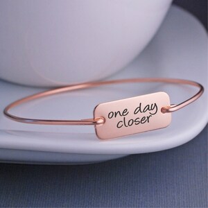 May include: A rose gold bangle bracelet with a rectangular charm that reads "one day closer".