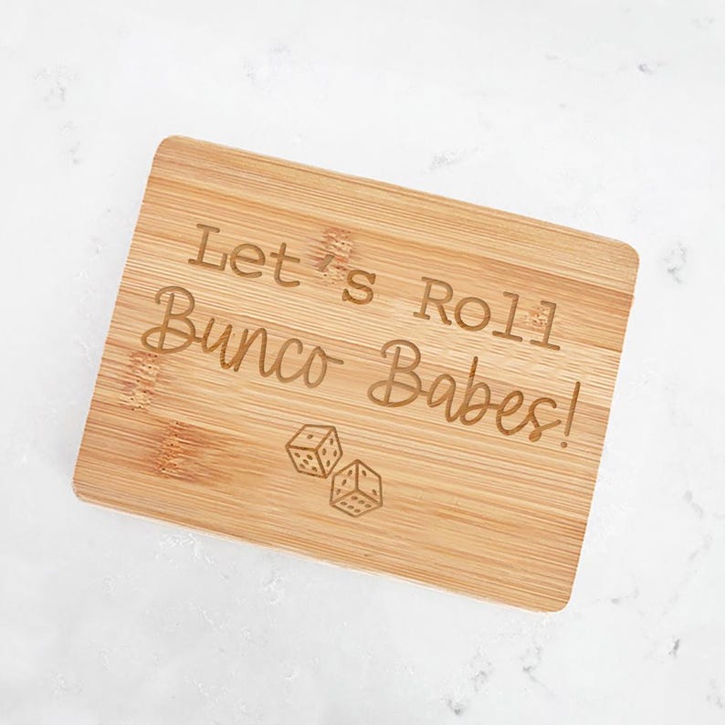 May include: A wooden cutting board with the text "Let's Roll Bunco Babes!" engraved on it. Two dice are also engraved on the board.