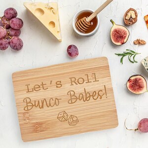 May include: A wooden cutting board with the text "Let's Roll Bunco Babes!" engraved on it. Two dice are also engraved on the board.