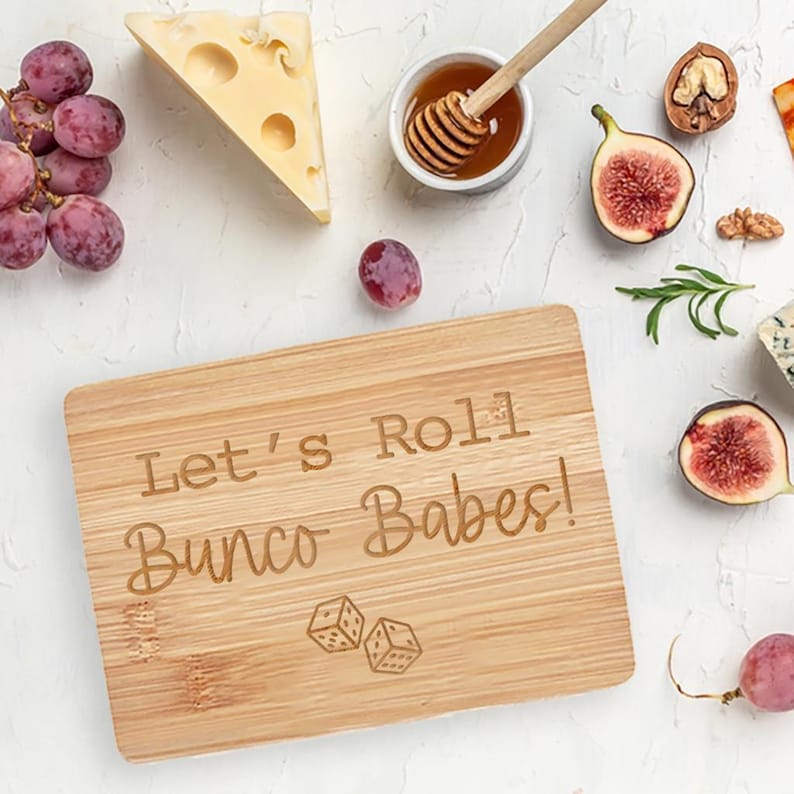 May include: A wooden cutting board with the text "Let's Roll Bunco Babes!" engraved on it. Two dice are also engraved on the board.