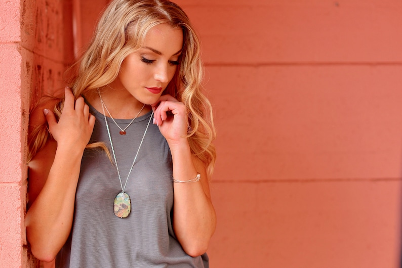 May include: A woman wearing a gray tank top with a silver necklace featuring a large, iridescent, oval pendant. She is standing in front of a peach-colored wall.
