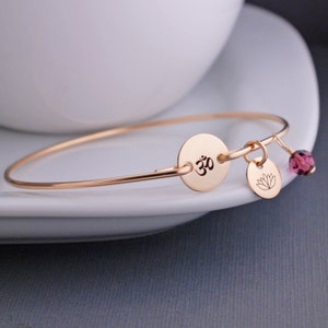 Puede incluir: Un brazalete de oro rosa con un dije que presenta el símbolo Om y una flor de loto. Una pequeña cuenta de cristal morado cuelga del dije.