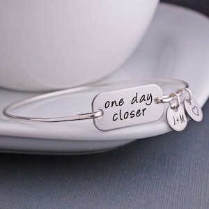 May include: Silver bangle bracelet with a charm that reads "one day closer". The bracelet also has two additional charms, one with the initials "J+M" and the other a heart.