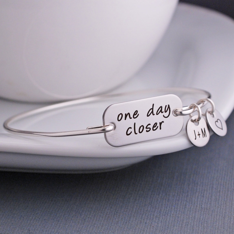 May include: Silver bangle bracelet with a charm that reads "one day closer". The bracelet also has two additional charms, one with the initials "J+M" and the other a heart.