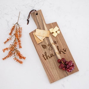 Może przedstawiać: Drewniana deska do serwowania z wygrawerowanym tekstem "give thanks". Deska jest ozdobiona winogronami, kostkami sera i krakersami.